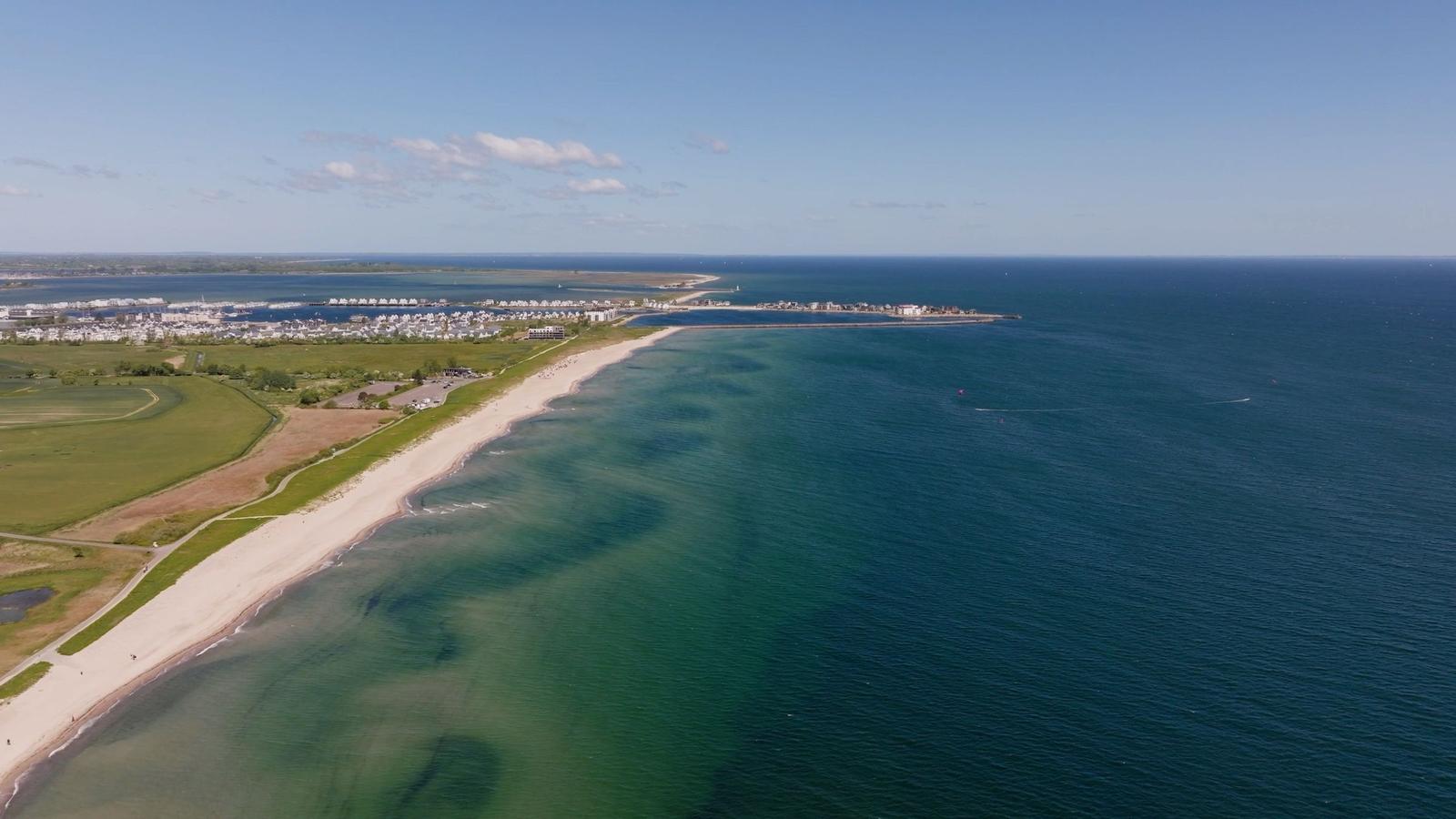 Strand mit Sandstrand, grünen Flächen und Hafen im Hintergrund.