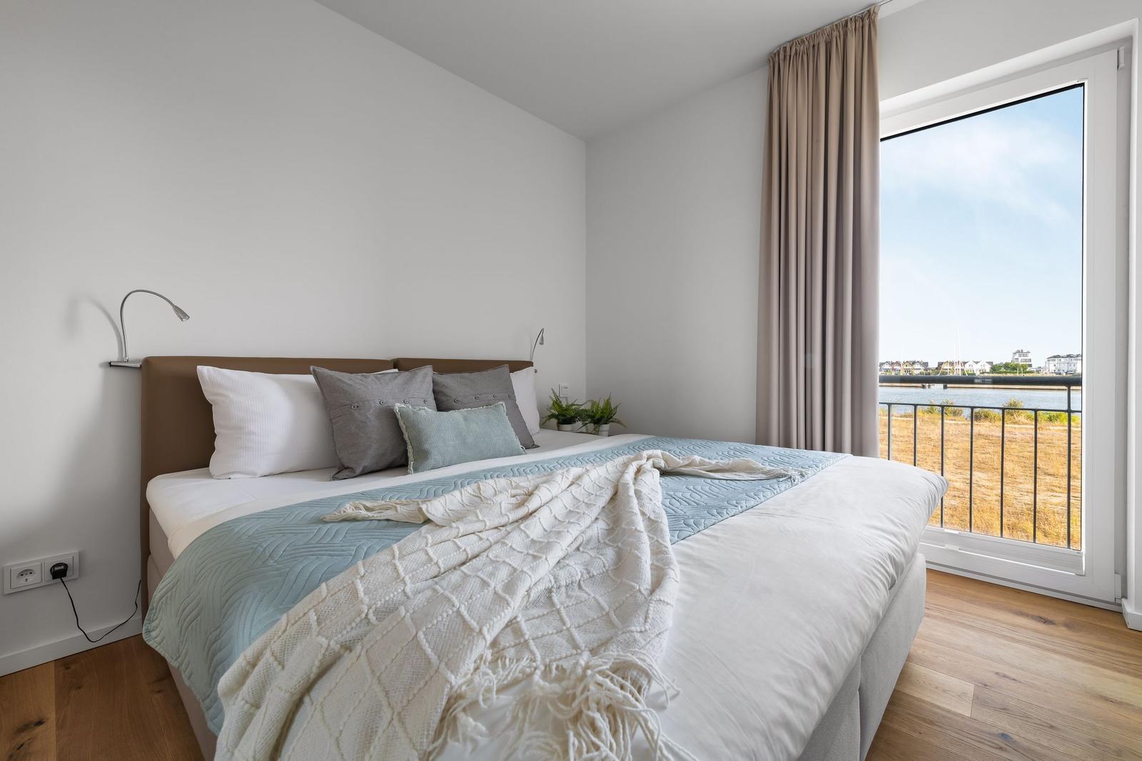 Schlafzimmer mit Blick auf Wasser, großes Bett mit Kissen und Decke, Holzboden und Fenster.