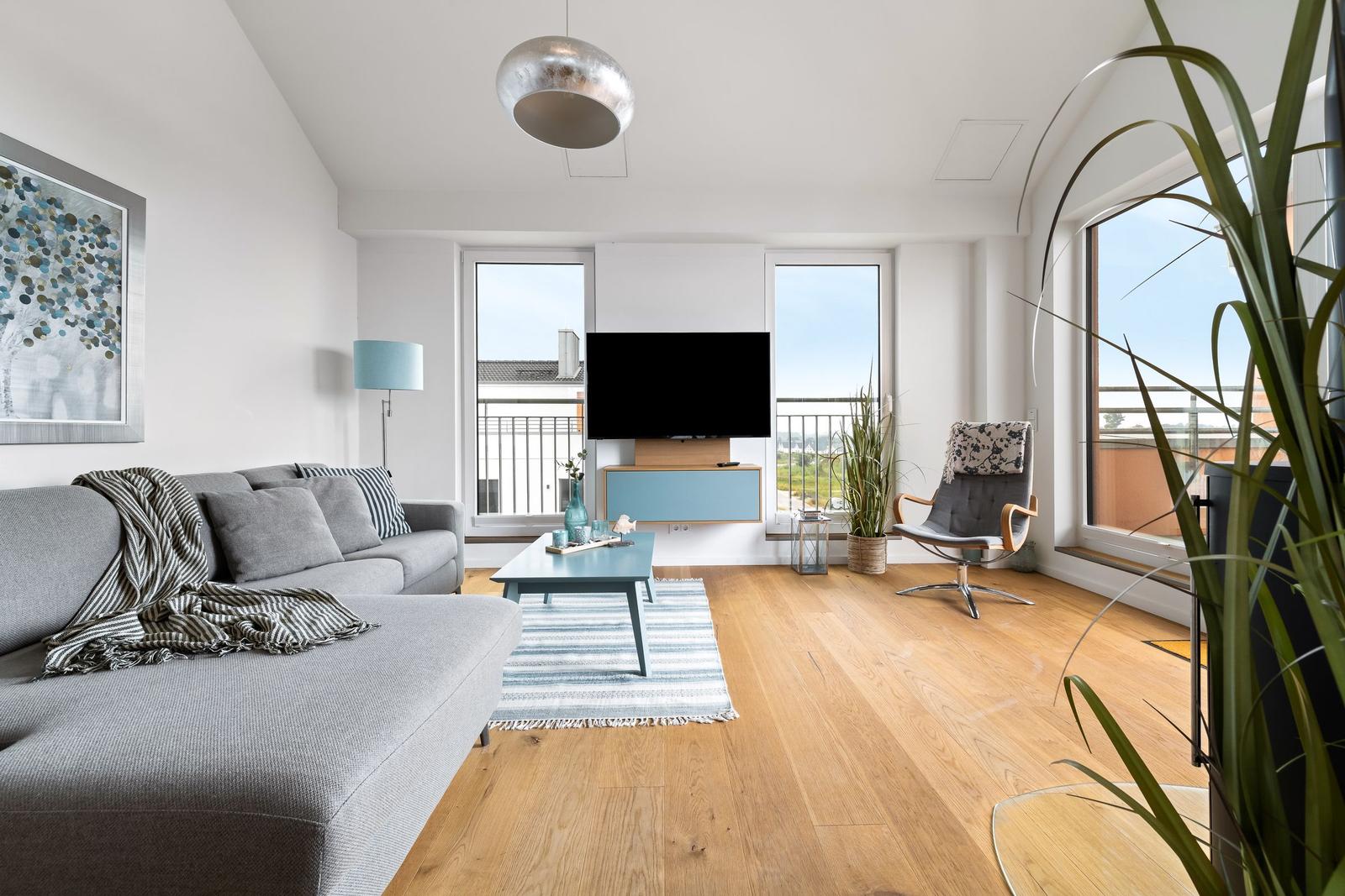 Wohnzimmer mit Sofa, Fernseher und Holzboden. Große Fenster und Pflanzen sorgen für viel Licht.