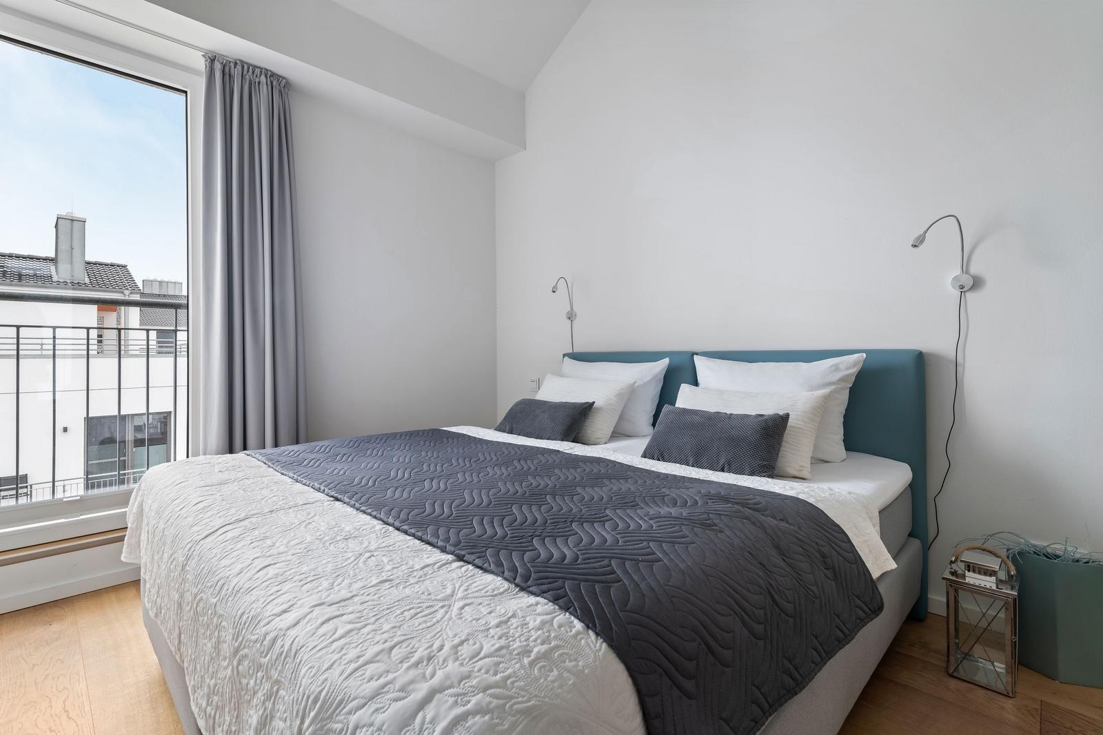 Schlafzimmer mit Bett, Fenster und Balkon. Wandlampen und graue Vorhänge.