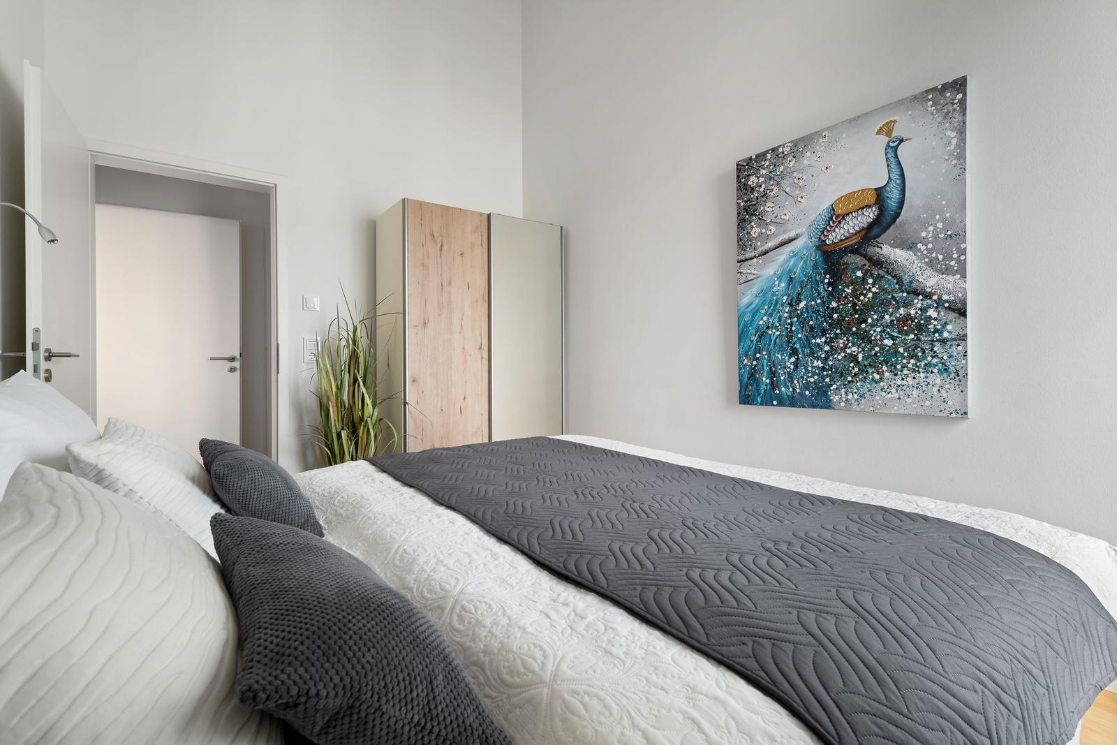 Schlafzimmer mit Bett, Kleiderschrank und Wandbild eines Pfauens.