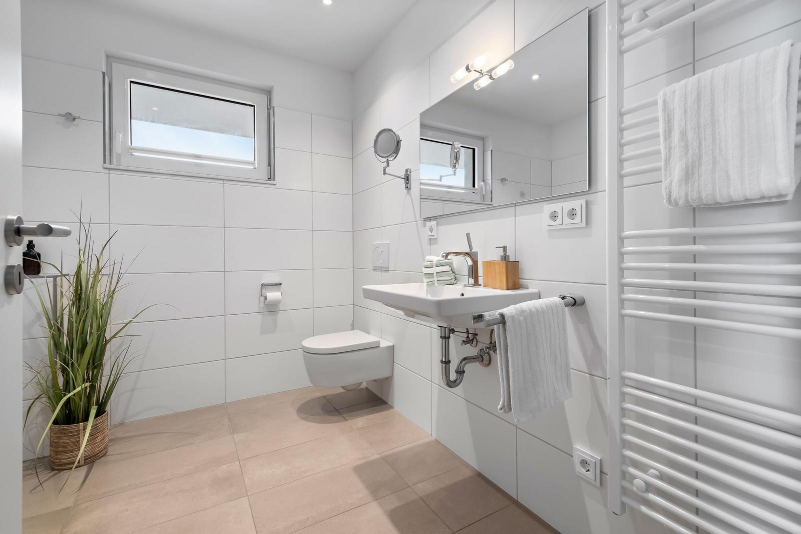 Badezimmer mit Toilette, Waschbecken und Heizkörper. Fenster, Spiegel und Pflanze sichtbar.