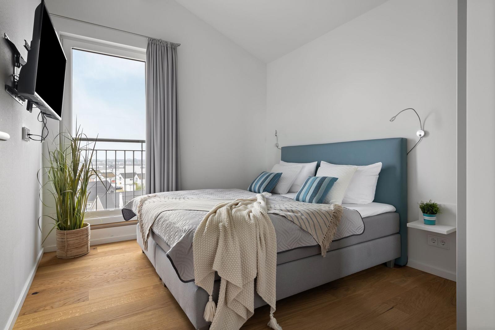 Schlafzimmer mit Bett, Fenster und Balkon. Wandmontierte TV und Pflanzen.