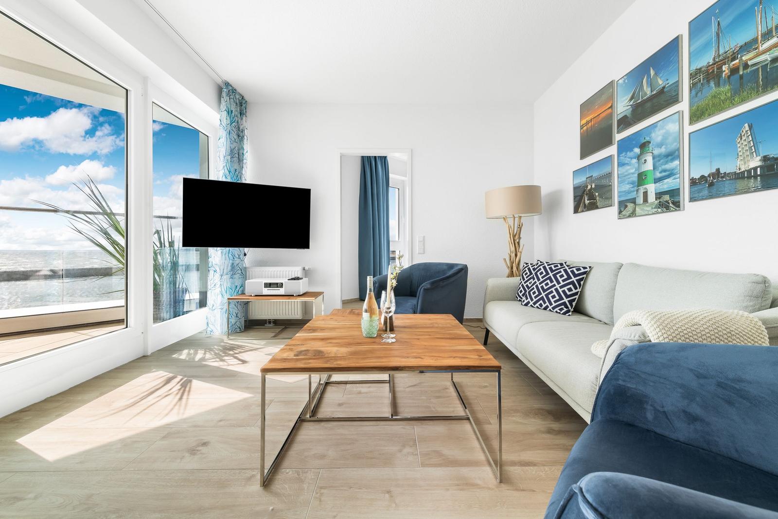 Wohnzimmer mit Sofa, Couchtisch und Fernseher. Große Fenster mit Meerblick.