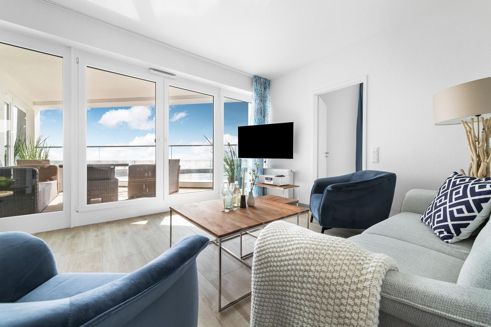 Wohnzimmer mit Blick auf Balkon und Meer. Sofa, Sessel und TV sichtbar.