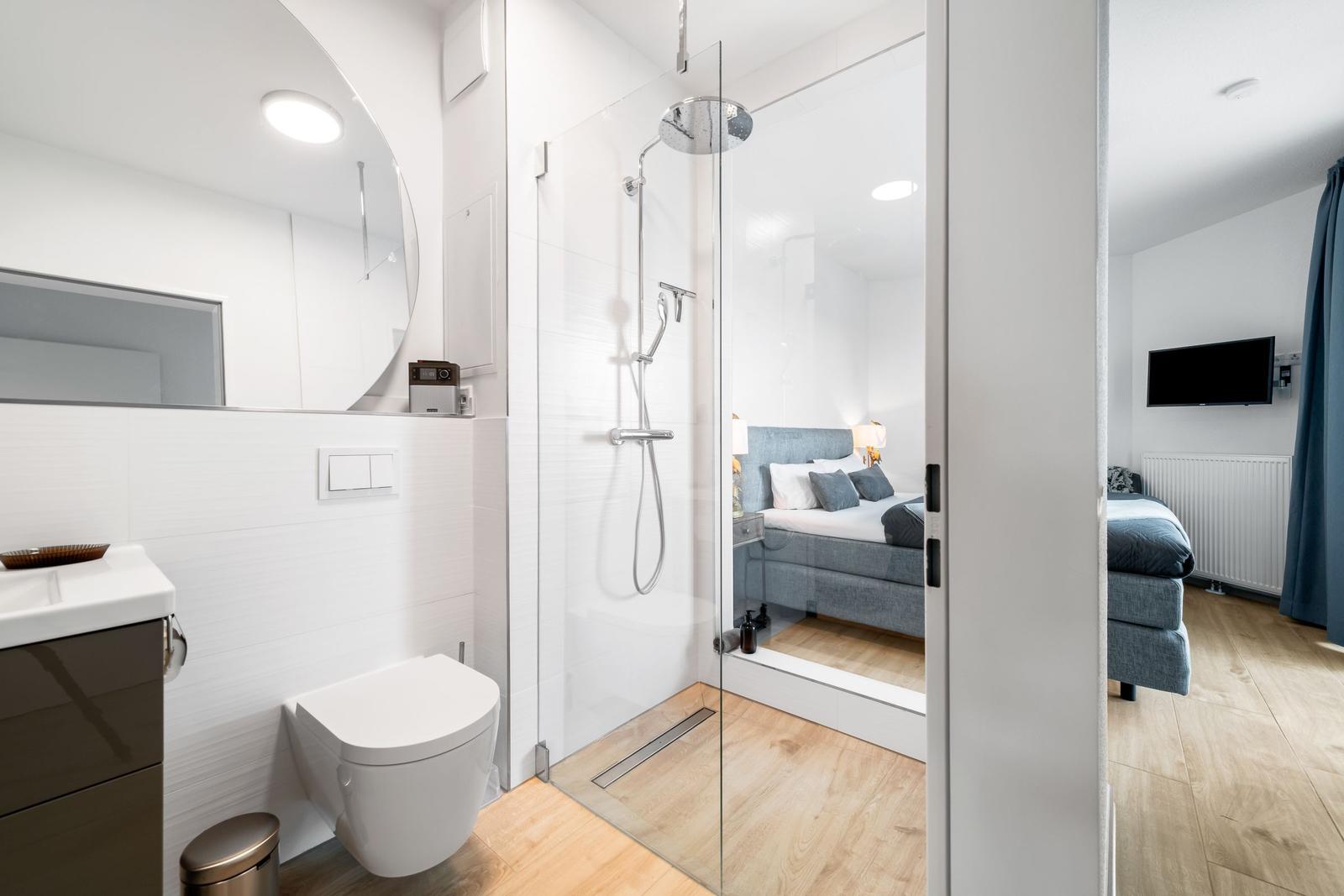 Badezimmer mit Dusche und Toilette. Im Hintergrund das Schlafzimmer mit Bett und Fernseher.