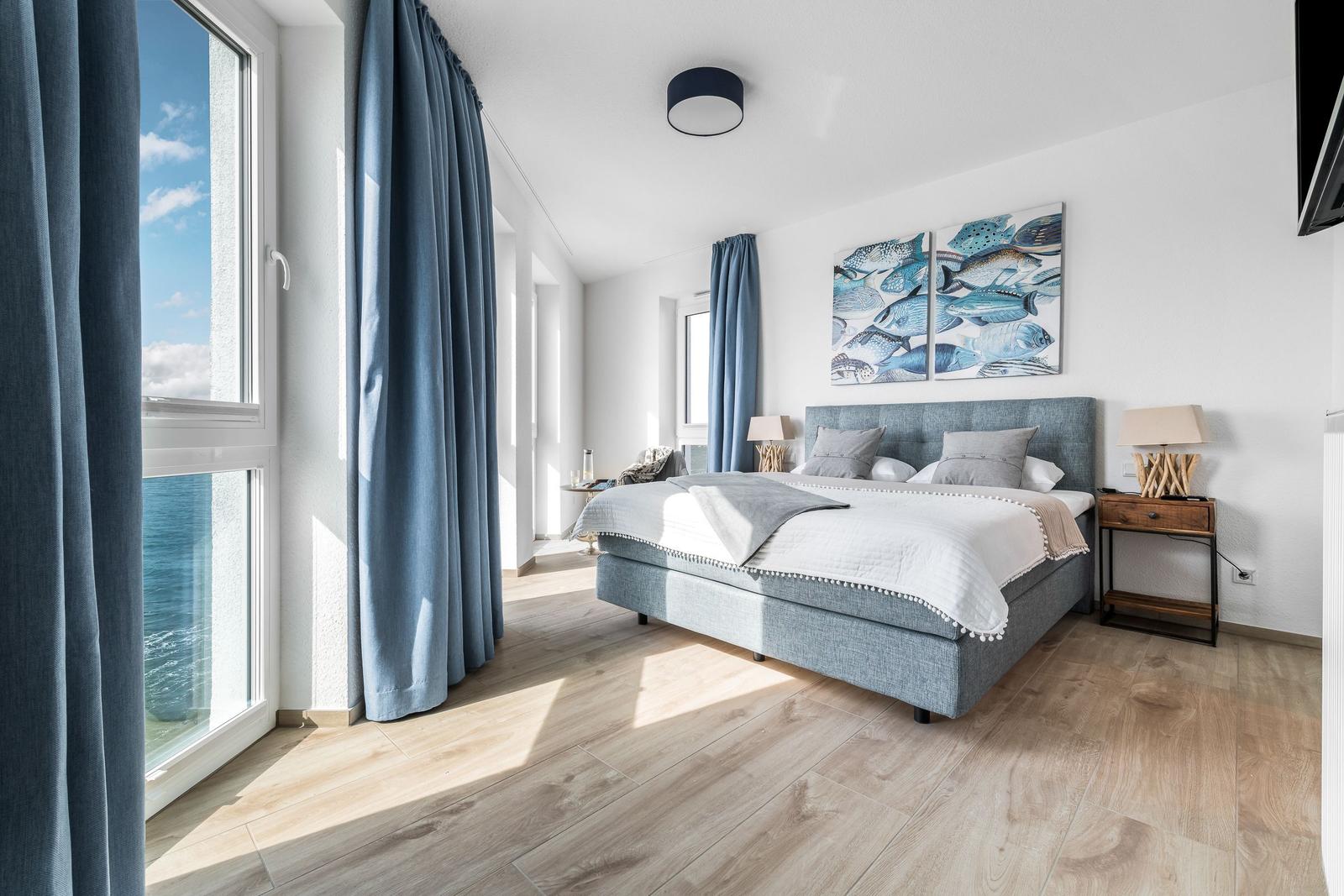 Schlafzimmer mit Blick aufs Meer, blauer Bettwäsche und Wandbild mit Fischen.