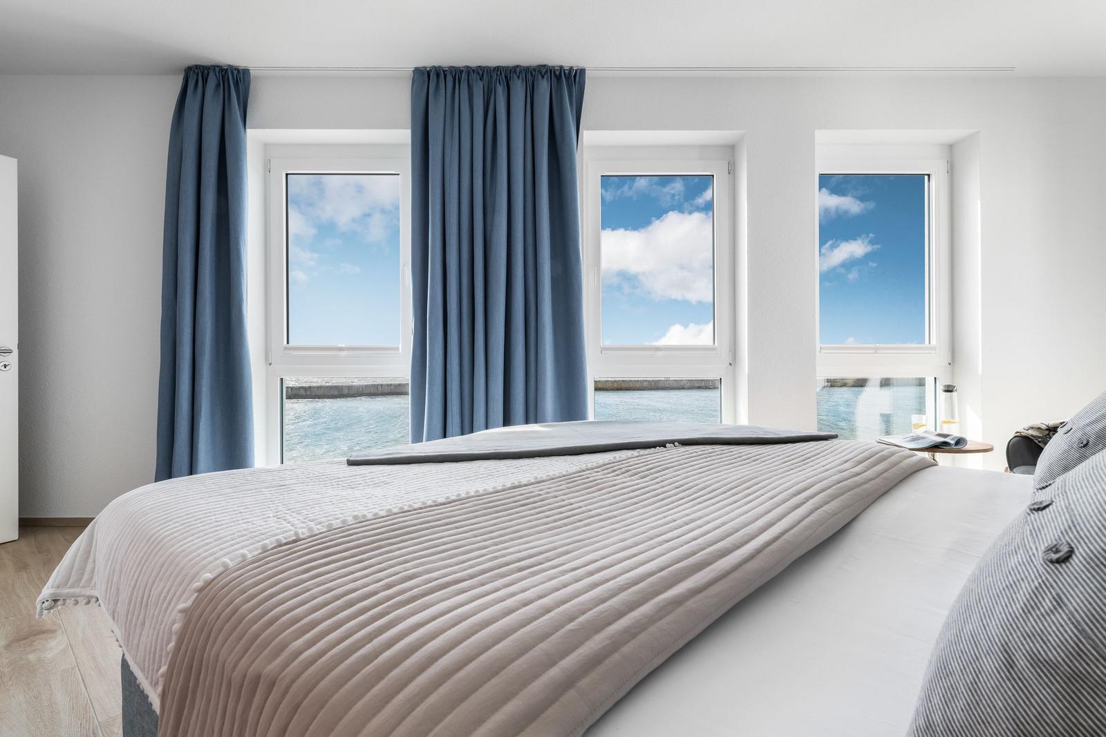 Schlafzimmer mit Blick aufs Meer durch drei Fenster. Bett mit gestreifter Bettdecke.