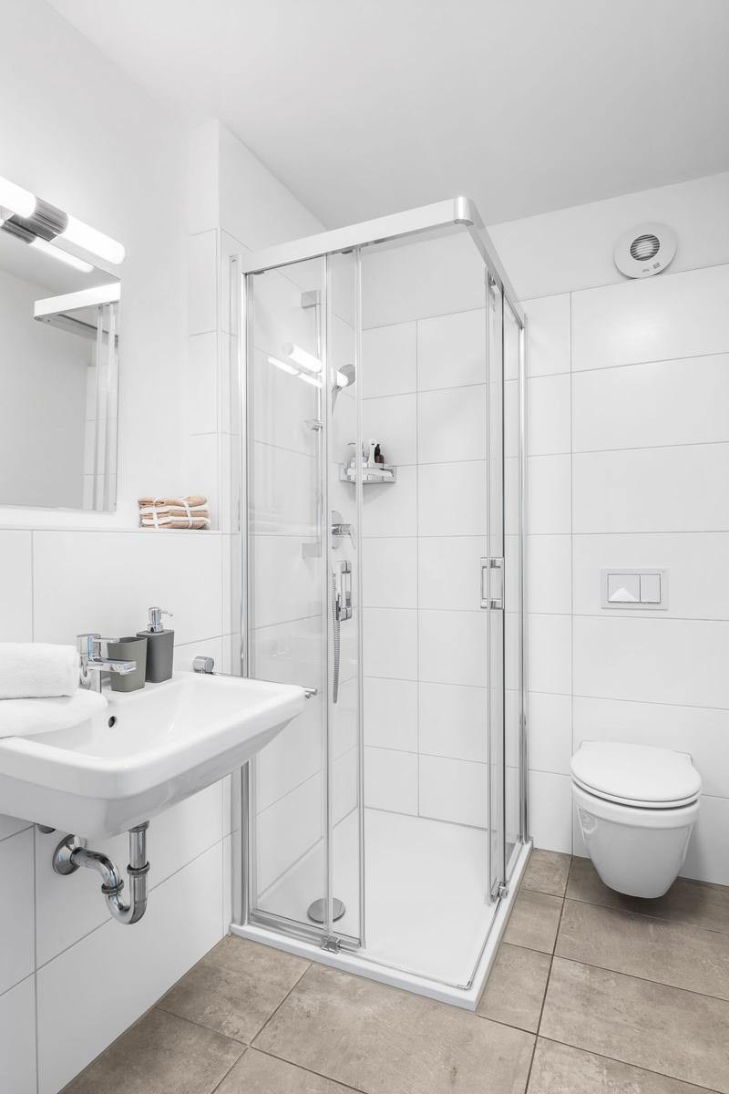 Badezimmer mit Dusche, Waschbecken und Toilette. Weiße Fliesen und bodenfarbener Fliesenboden.