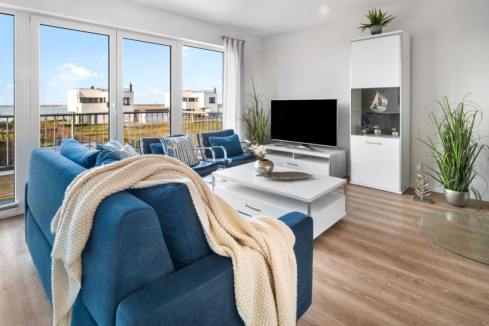 Wohnzimmer mit blauem Sofa, TV und Blick auf Balkon und Meer.