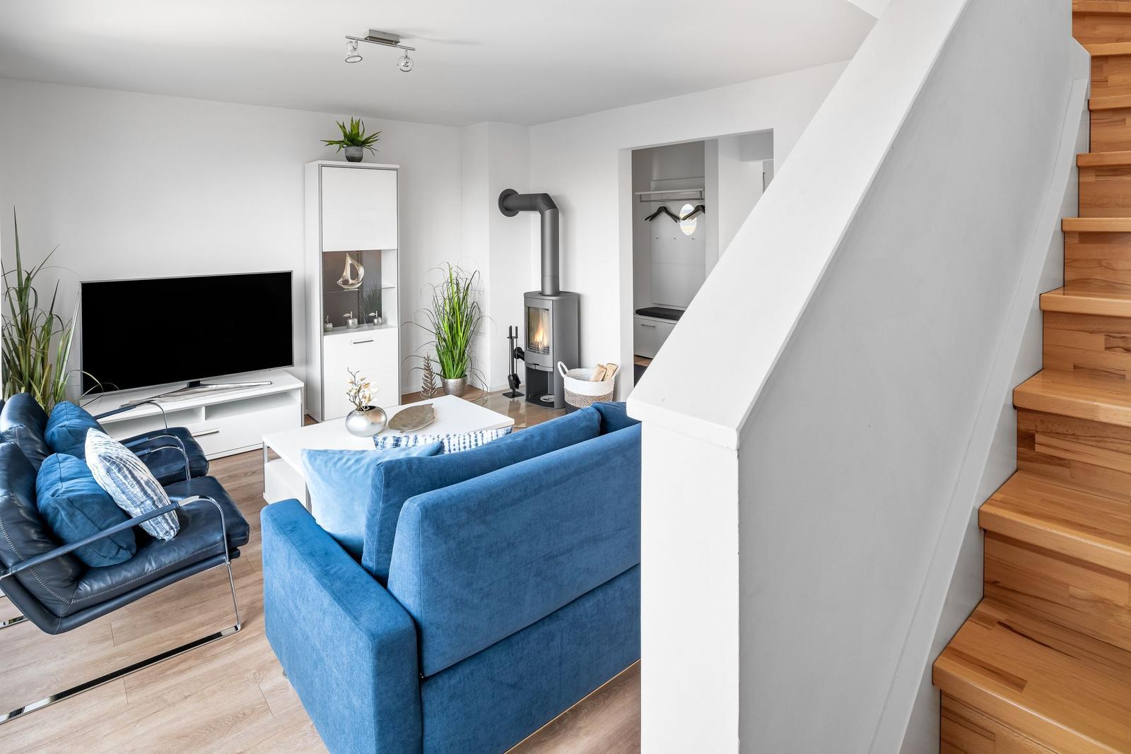Wohnzimmer mit blauem Sofa, Fernseher und Kaminofen. Holztreppe führt nach oben.