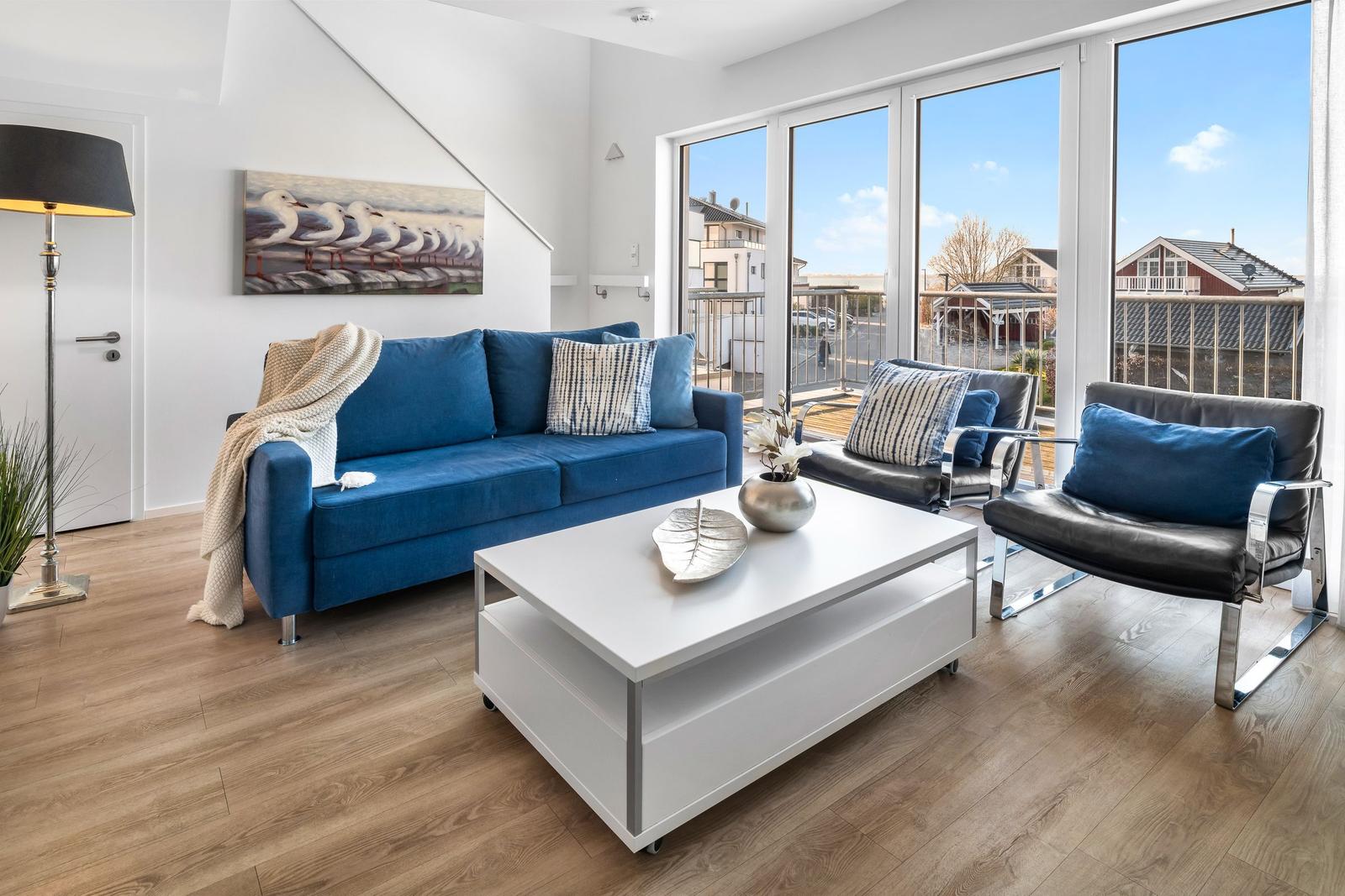 Ein Wohnzimmer mit blauem Sofa, schwarzen Sesseln und großen Fenstern zur Terrasse.