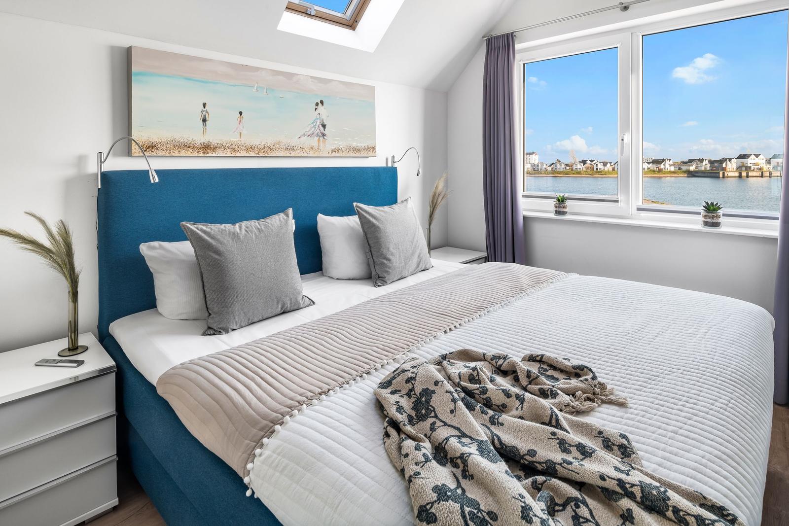 Schlafzimmer mit Blick auf Wasser und Häuser. Blauer Kopfteil, Fenster und Wandbild.