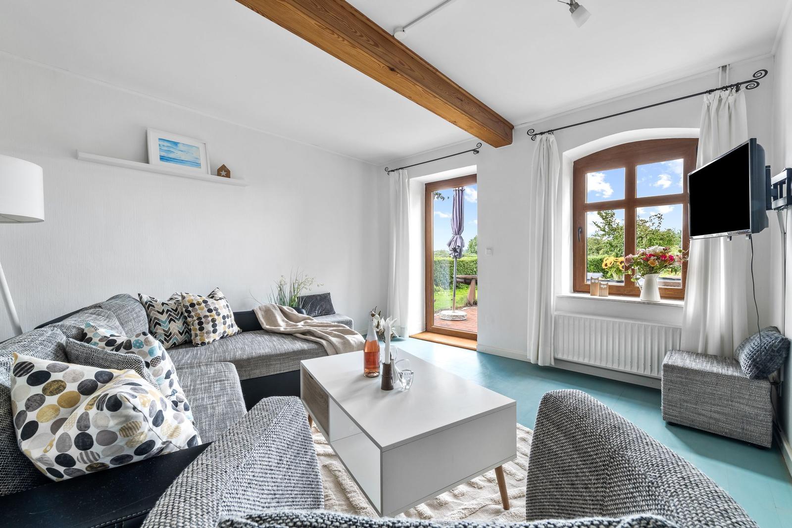 Wohnzimmer mit Sofa, Couchtisch und Fenster zur Terrasse. Holzbalken und blauer Boden.