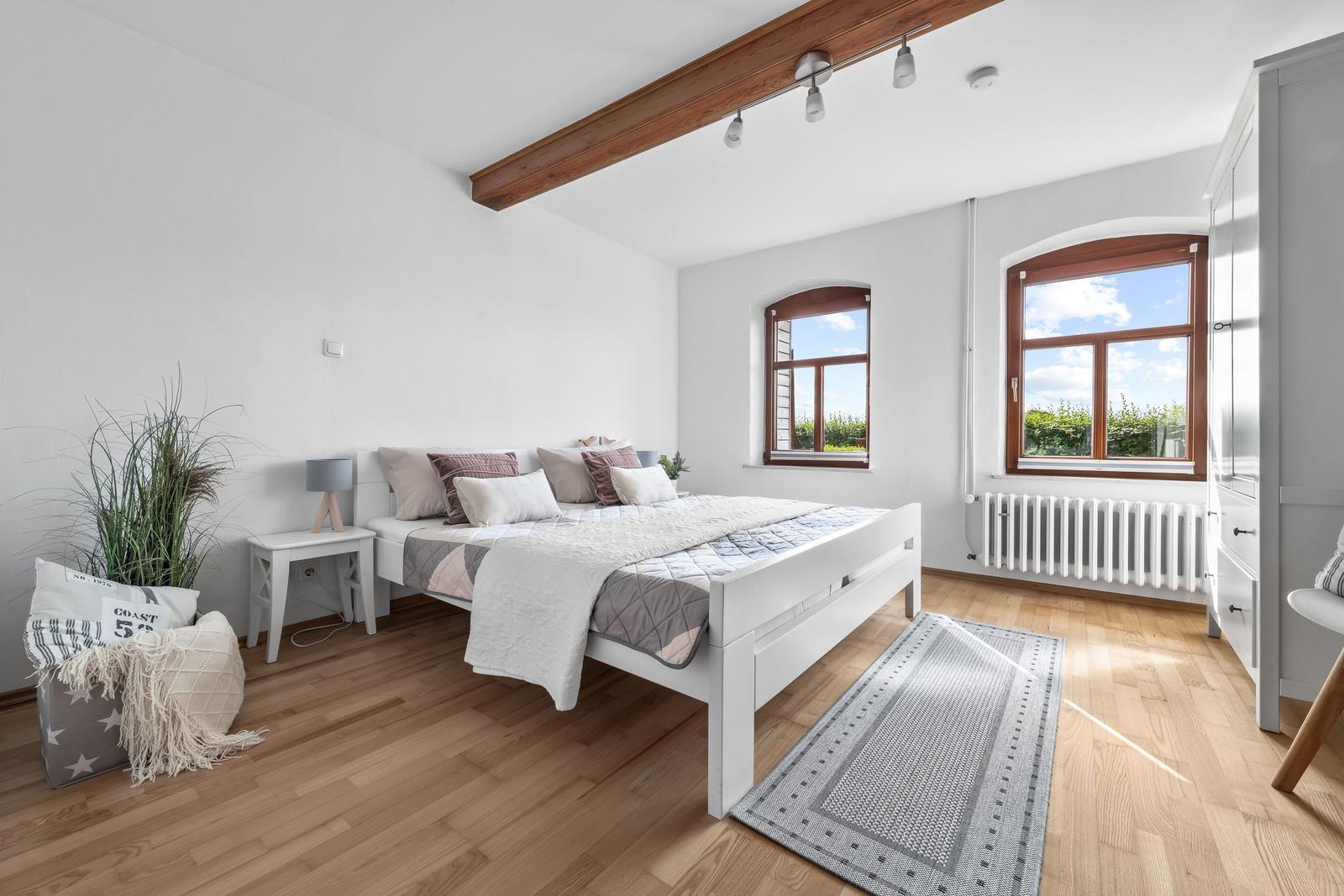 Schlafzimmer mit weißem Bett, Holzboden und zwei Fenstern mit Blick nach draußen.