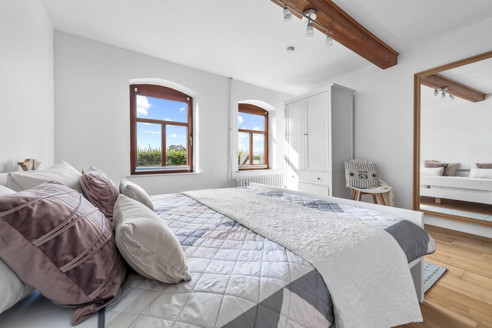 Schlafzimmer mit Doppelbett, zwei Fenstern und Spiegeltür. Holzbalken und Kleiderschrank sichtbar.