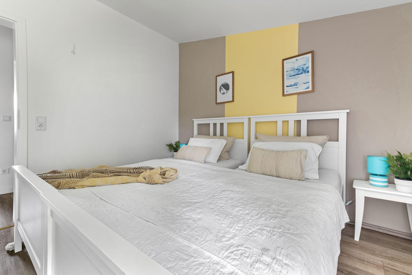 Schlafzimmer mit weißem Bett, gelb-brauner Wand und Nachttisch mit Lampe.
