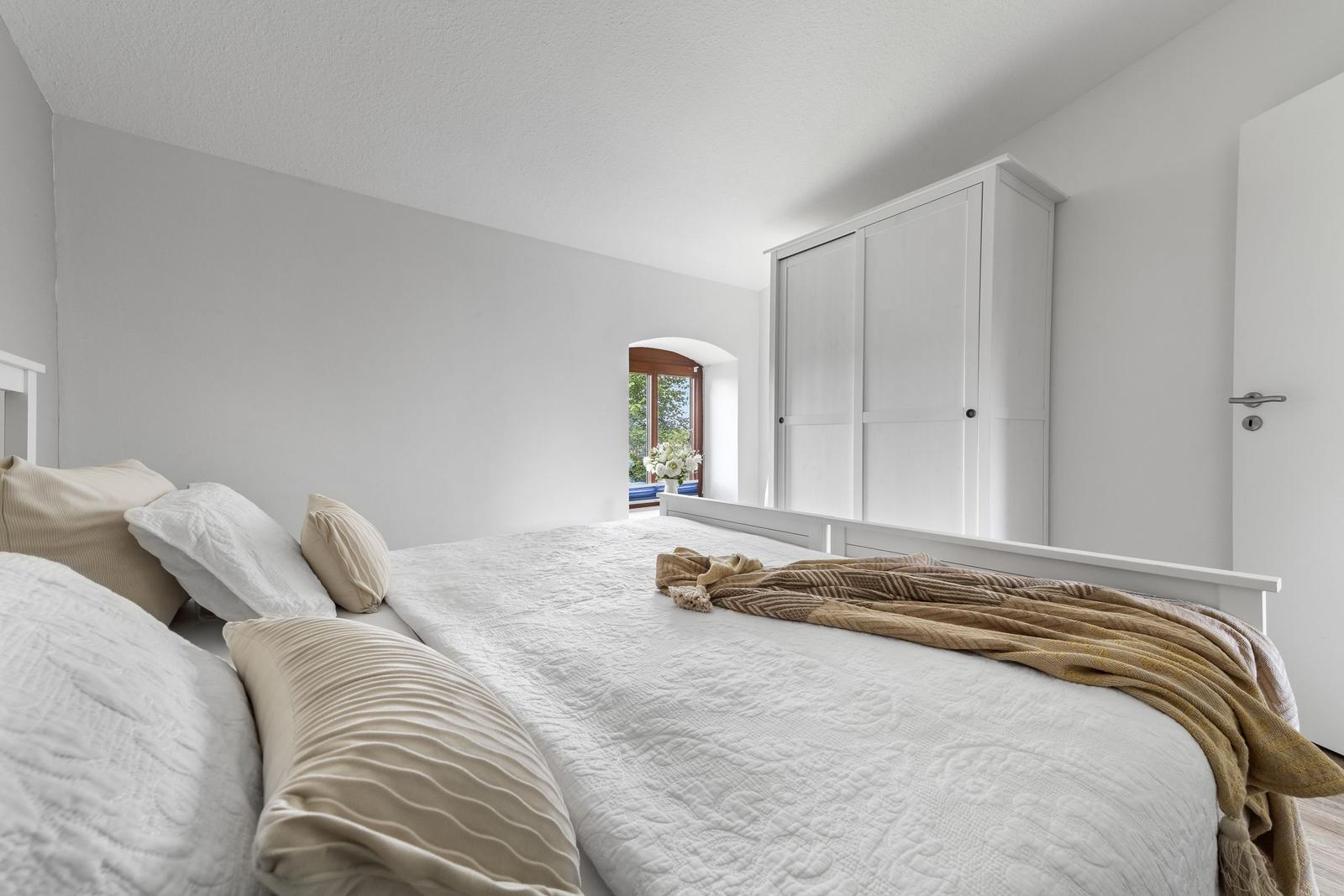 Schlafzimmer mit weißem Bett, Kleiderschrank und Fenster mit Blick nach draußen.