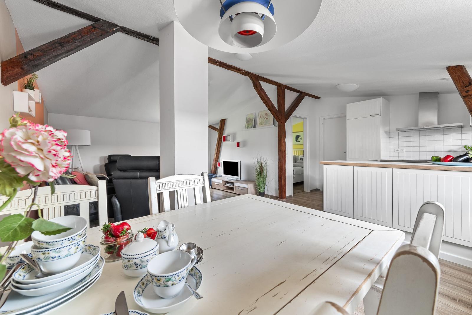 Essbereich mit Tisch und Stühlen. Küche und Wohnzimmer im Hintergrund. Holzbalken und weiße Möbel.