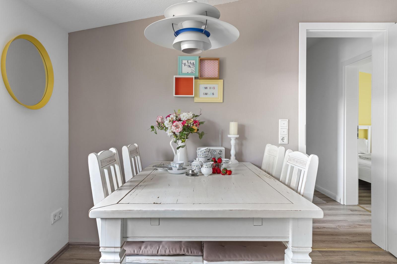 Esszimmer mit weißem Tisch, Stühlen und Deckenleuchte. Wand mit Bilderrahmen und Spiegel.