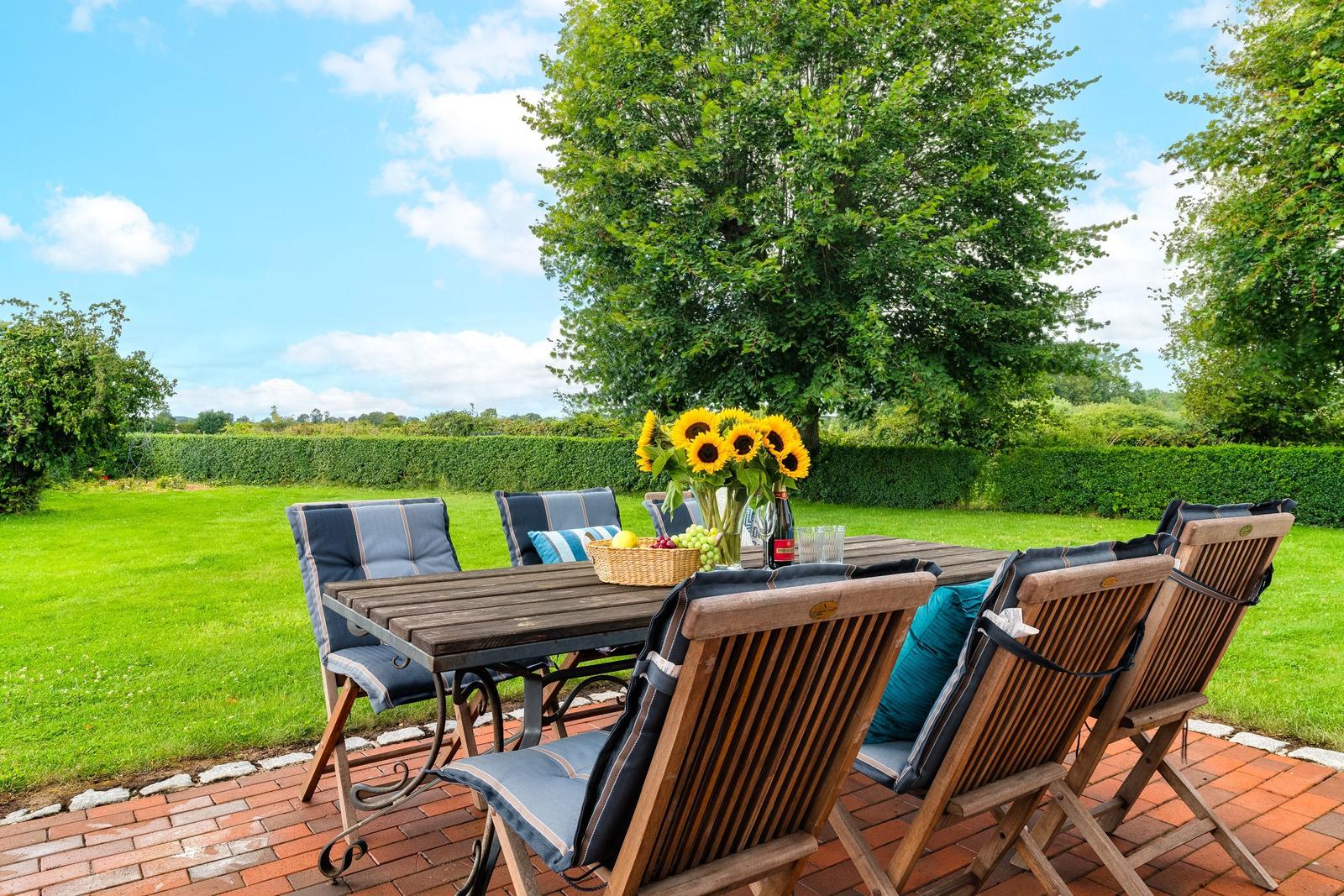 Holztisch mit Stühlen auf Terrasse. Sonnenblumen, Obst und Getränk darauf. Grünfläche und Bäume im Hintergrund.