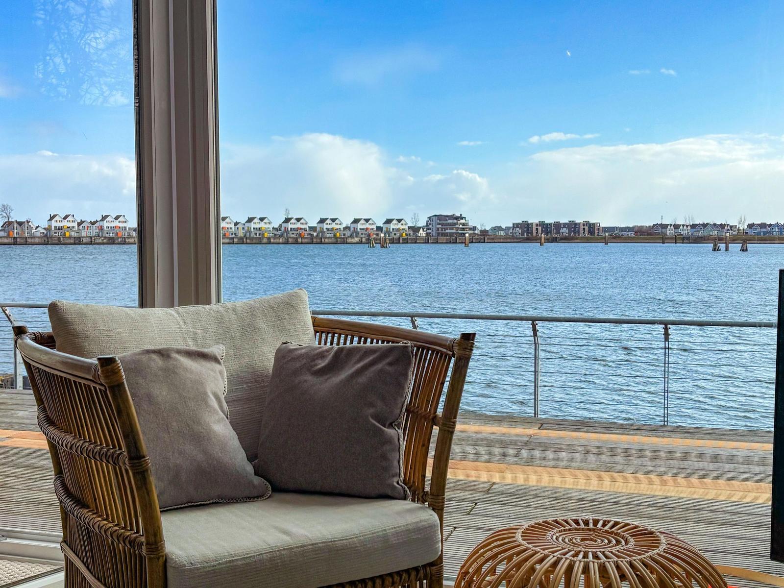 Rattan-Sofa mit Kissen auf Terrasse mit Blick auf Wasser und gegenüberliegende Gebäude.