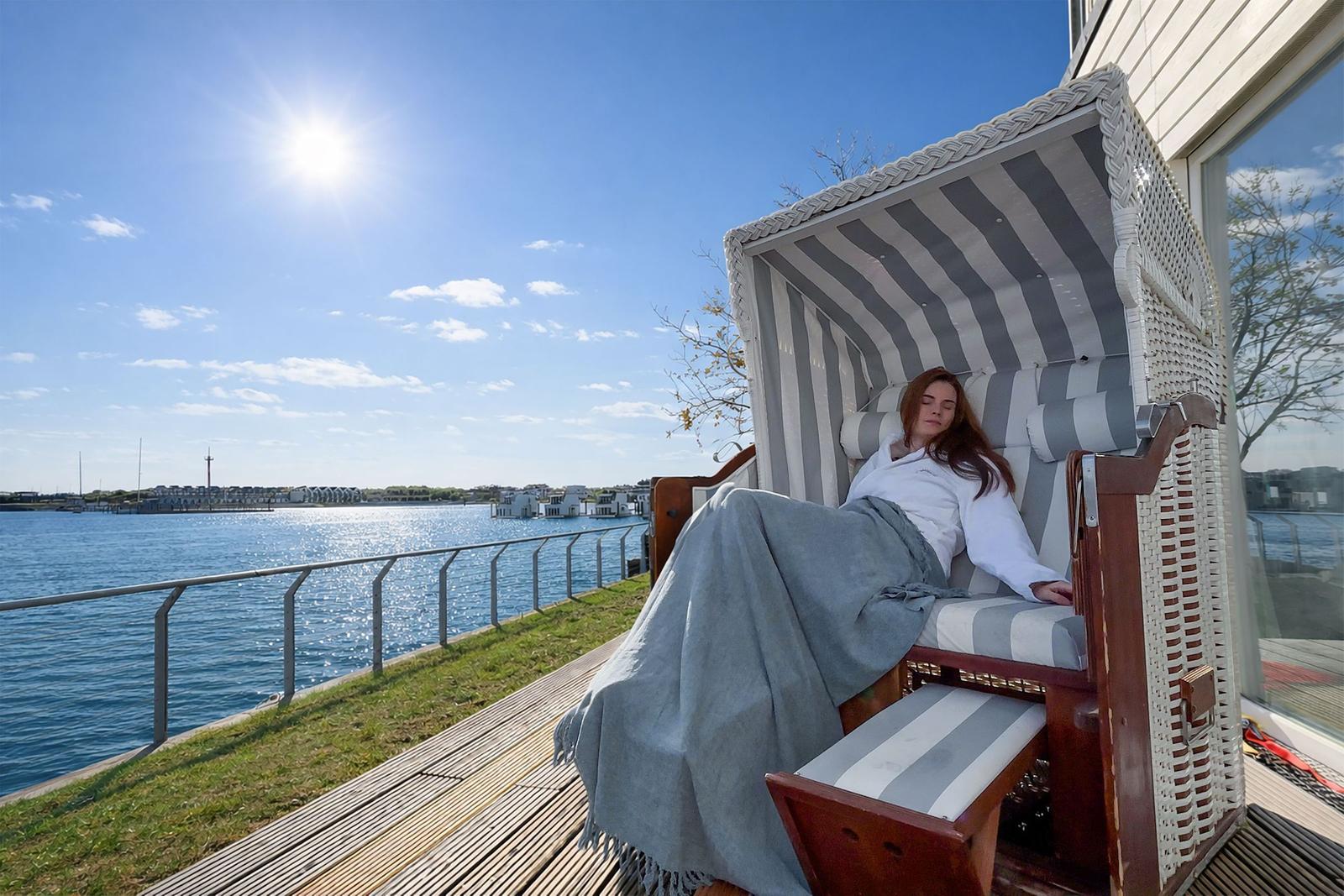 Eine Person ruht in einem Strandkorb auf einer Terrasse mit Blick auf Wasser und Himmel.