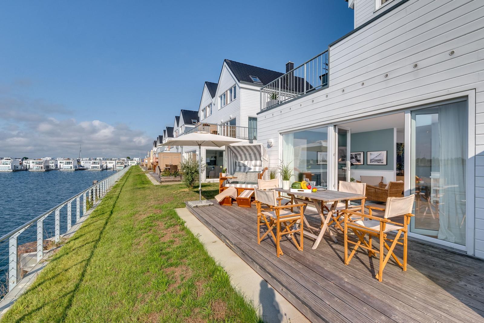Terrasse mit Tisch, Stühlen und Sonnenschirm neben Wasser. Modernes Haus mit Glasfront.