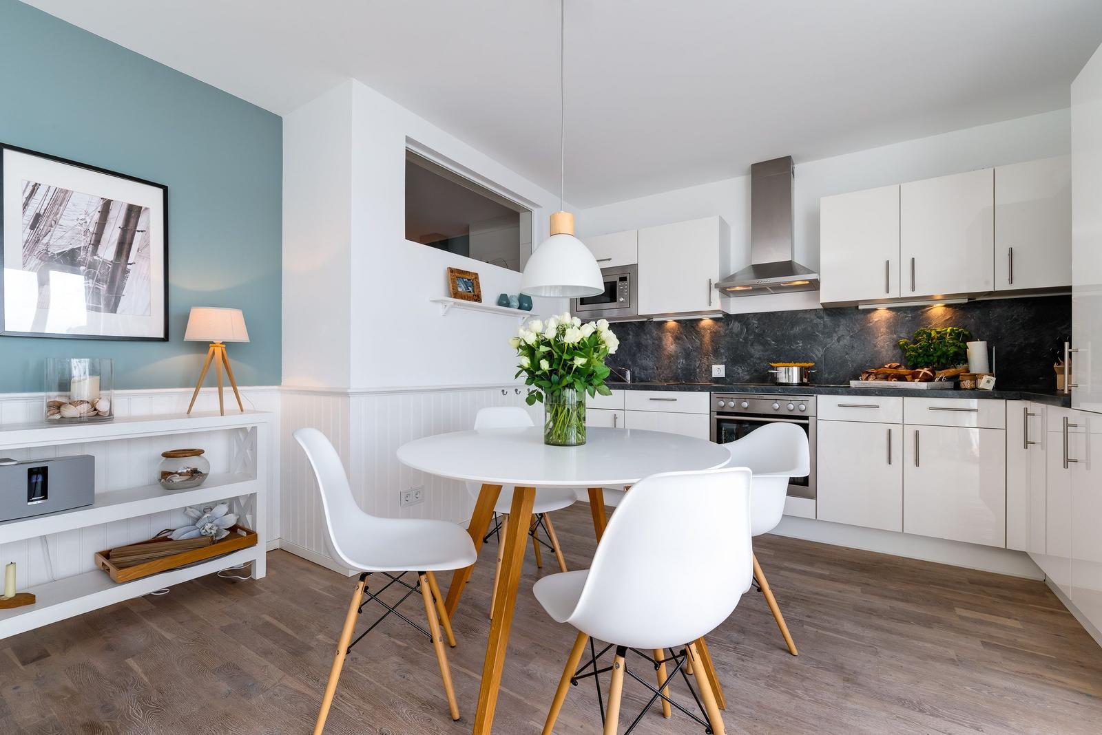 Küche mit Essbereich, weißer Tisch, vier Stühle, dunkler Arbeitsplatte, weiße Schränke.