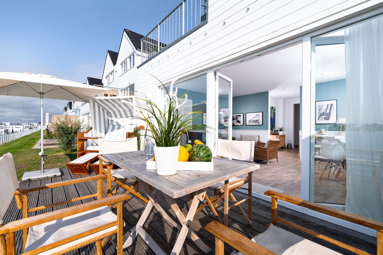Terrasse mit Tisch, Stühlen und Sonnenschirm. Durchgang in Wohnbereich mit Sofa und Wandbildern.