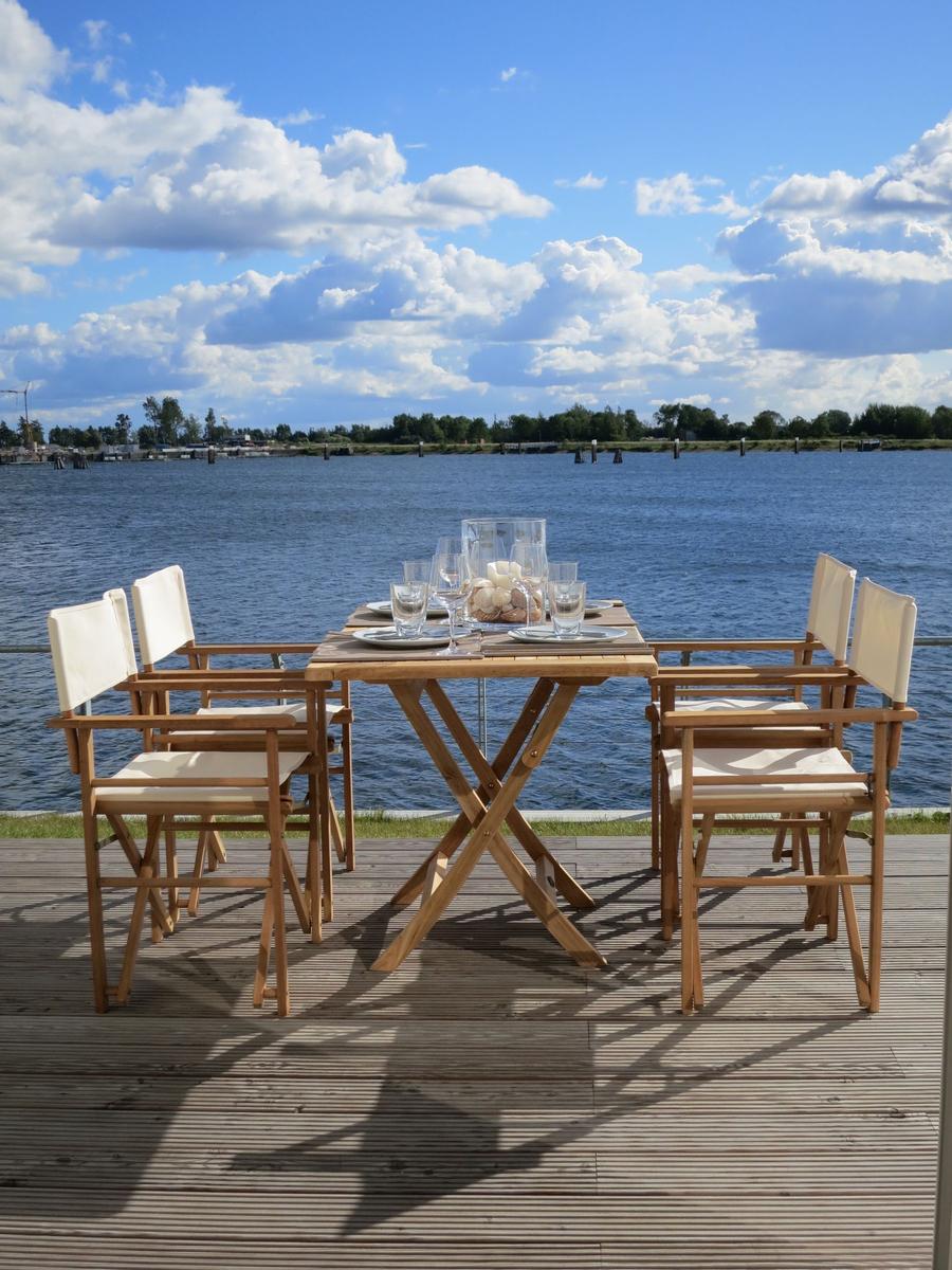 Holztisch mit Stühlen auf Terrasse mit Blick auf Wasser und Himmel.