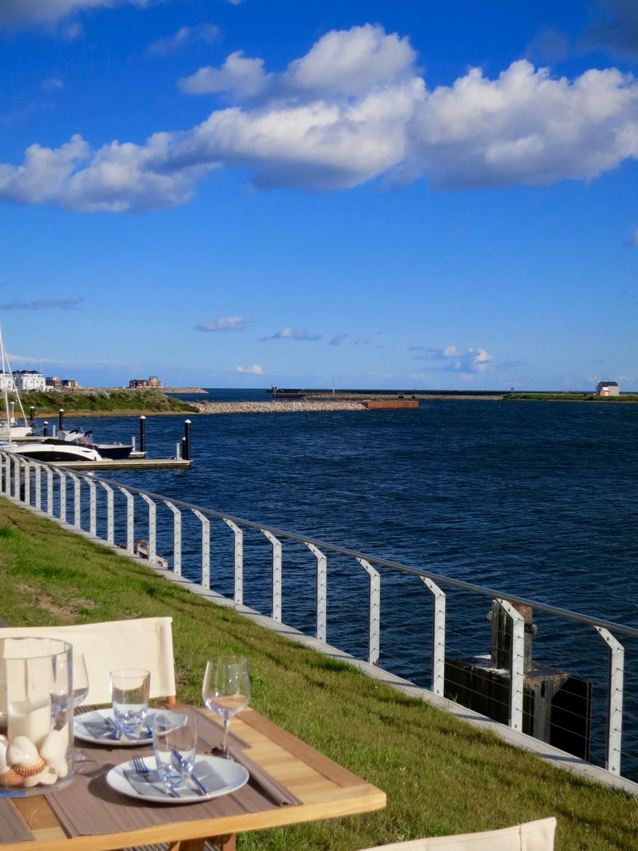 Tisch mit Besteck und Gläsern auf Grasfläche am Wasser mit Blick auf Hafen und blaues Meer.