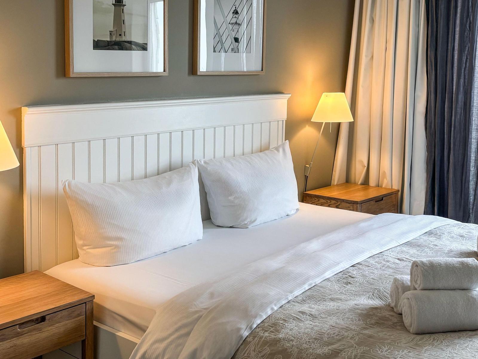Ein Bett mit weißen Kissen und einer Decke. Zwei Nachttische mit Lampen und gerollte Handtücher.