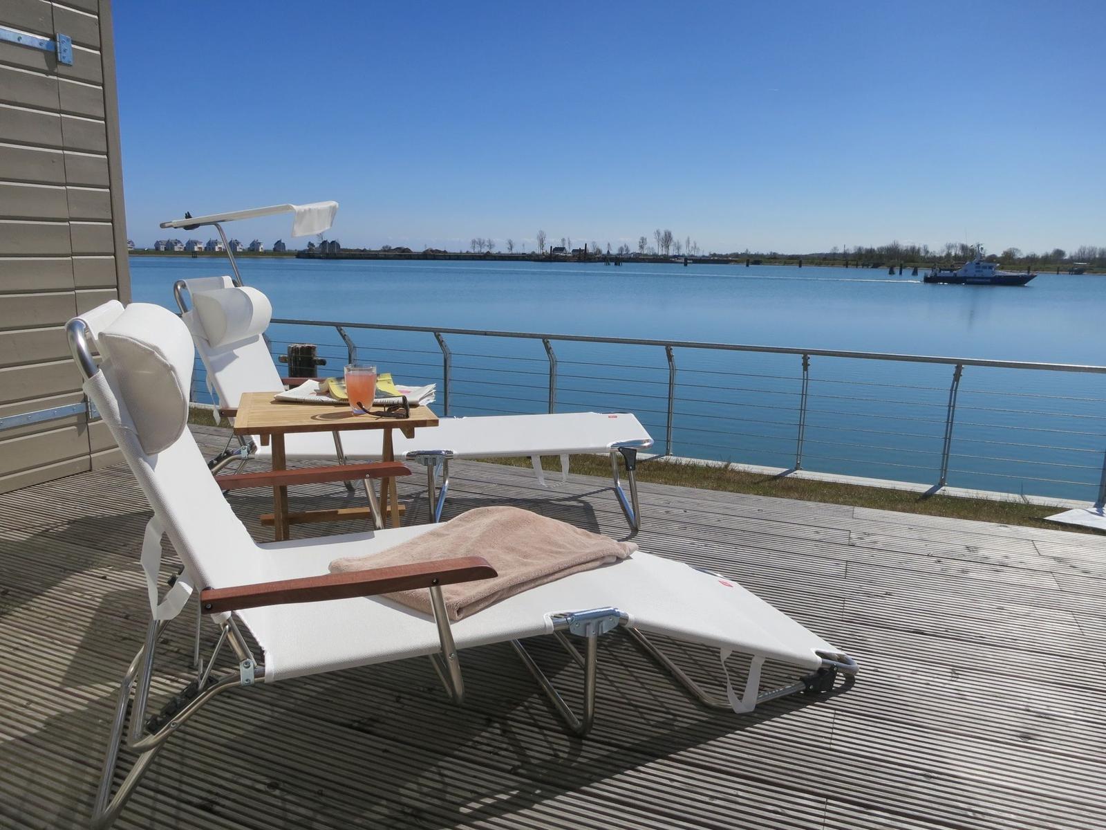 Terrasse mit Liegestühlen und Tisch am Wasser. Boot fährt im Hintergrund.