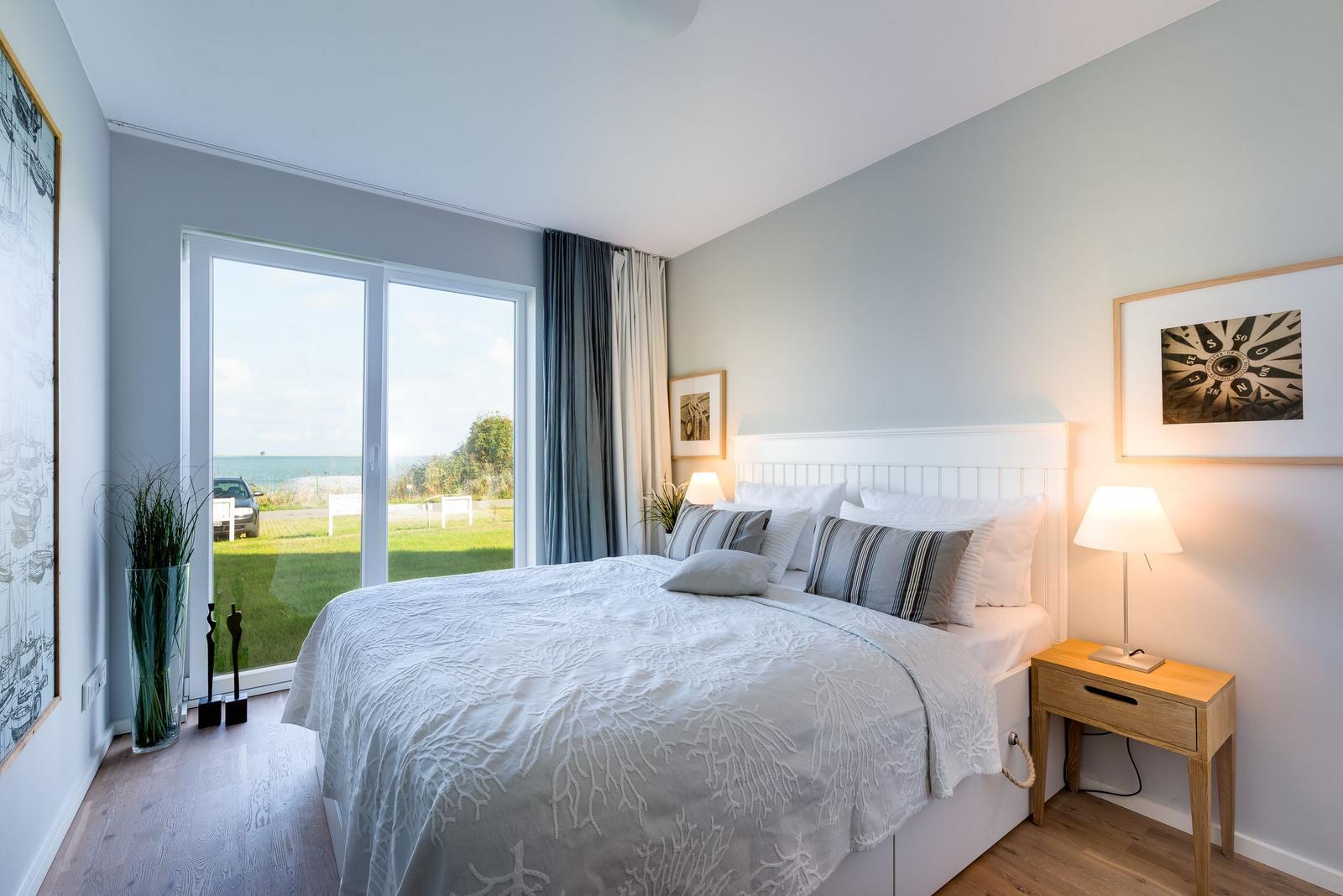 Schlafzimmer mit Blick auf den Strand und grünen Rasen. Bett, Nachttisch und Fenster sind sichtbar.