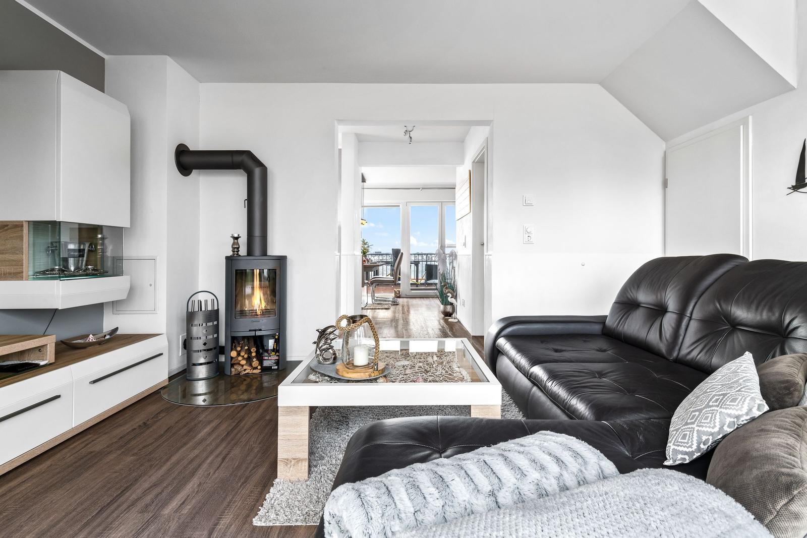 Wohnzimmer mit schwarzer Ledercouch, Holzboden und Kaminofen. Durchgang zum Balkon mit Blick.