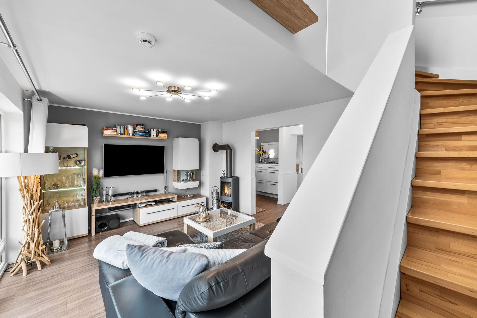 Wohnzimmer mit Sofa, Fernseher und Kamin. Holztreppe führt zur oberen Etage.