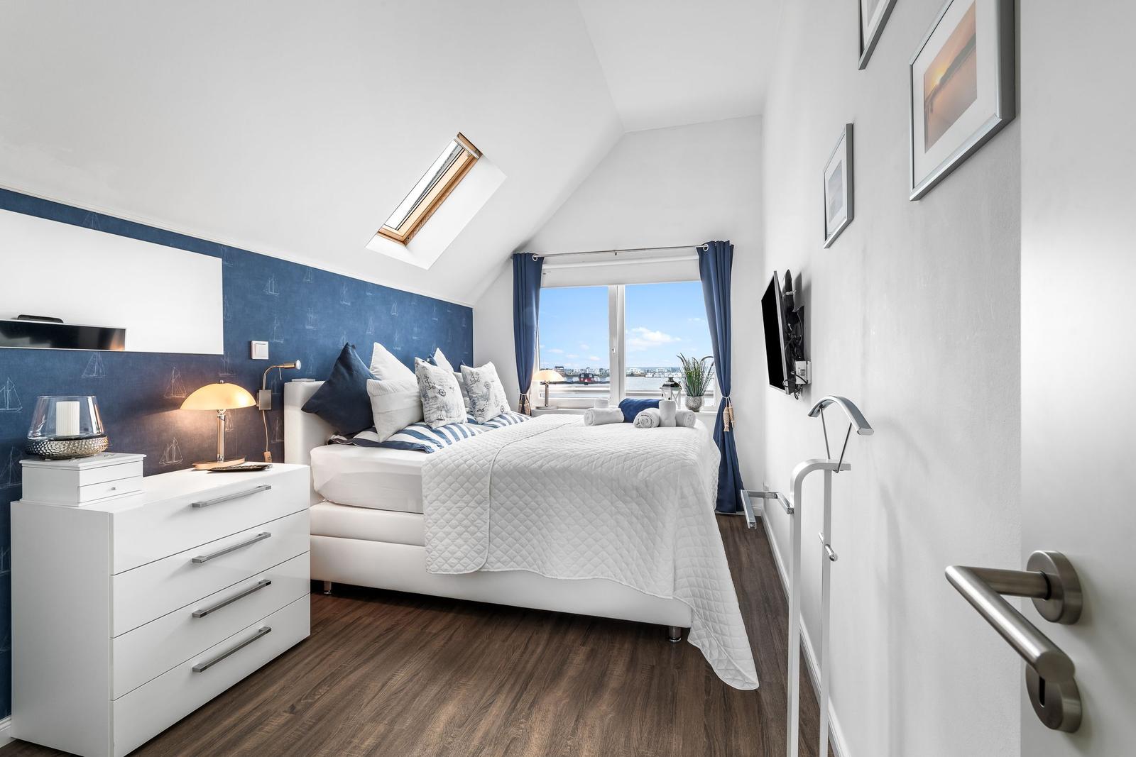 Schlafzimmer mit Blick auf Wasser, weißes Bett, blaue Wand und Dachschrägen.