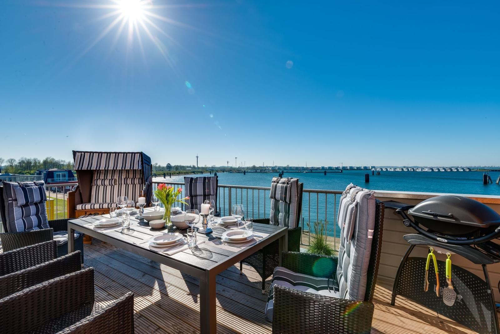 Terrasse mit Tisch, Stühlen und Grill. Blick auf Wasser und Himmel.