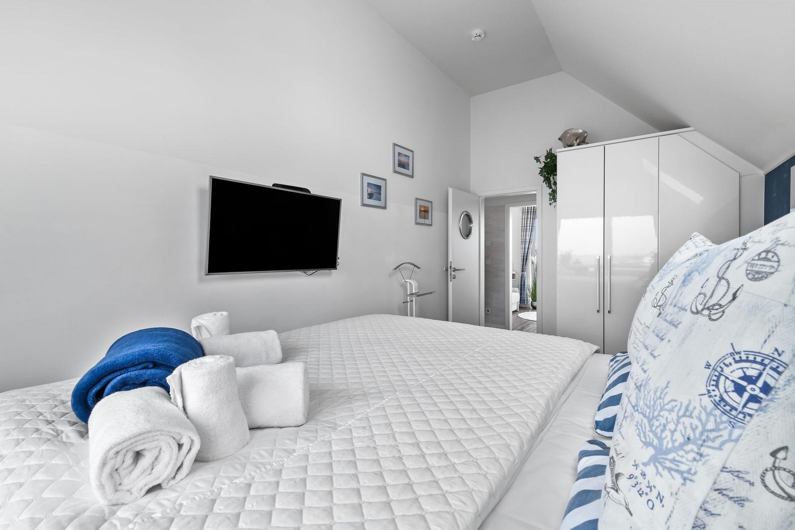 Schlafzimmer mit Bett, TV und Kleiderschrank. Handtuchrollen und maritimes Kissen.