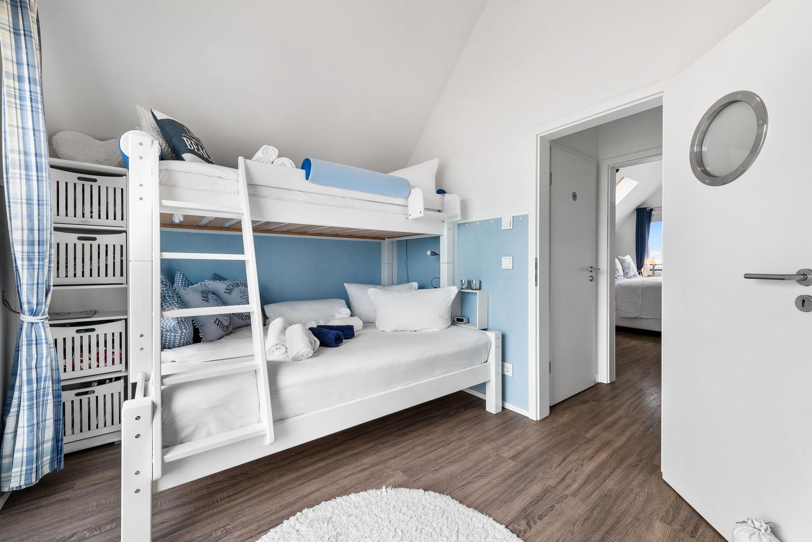 Zimmer mit Doppelstockbett, blauer Wand und Holzboden. Tür führt in ein weiteres Zimmer.