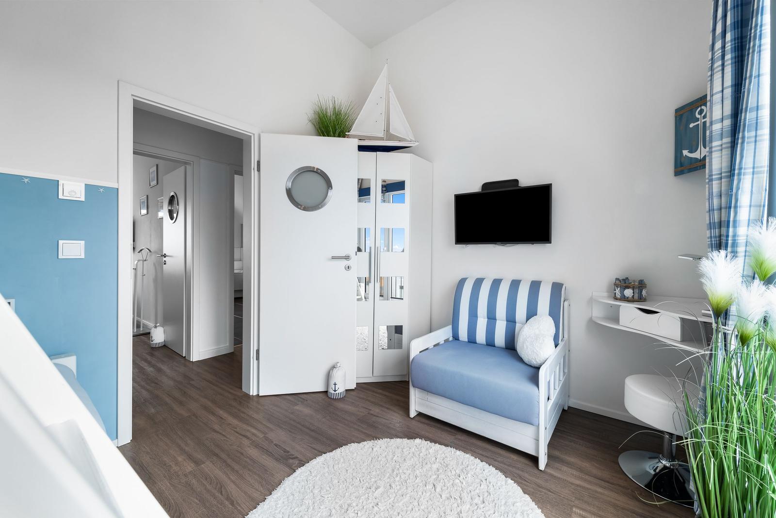 Kinderzimmer mit blau-weißem Sofa, TV und Schreibtisch. An Decke hängt ein Segelbootmodell.
