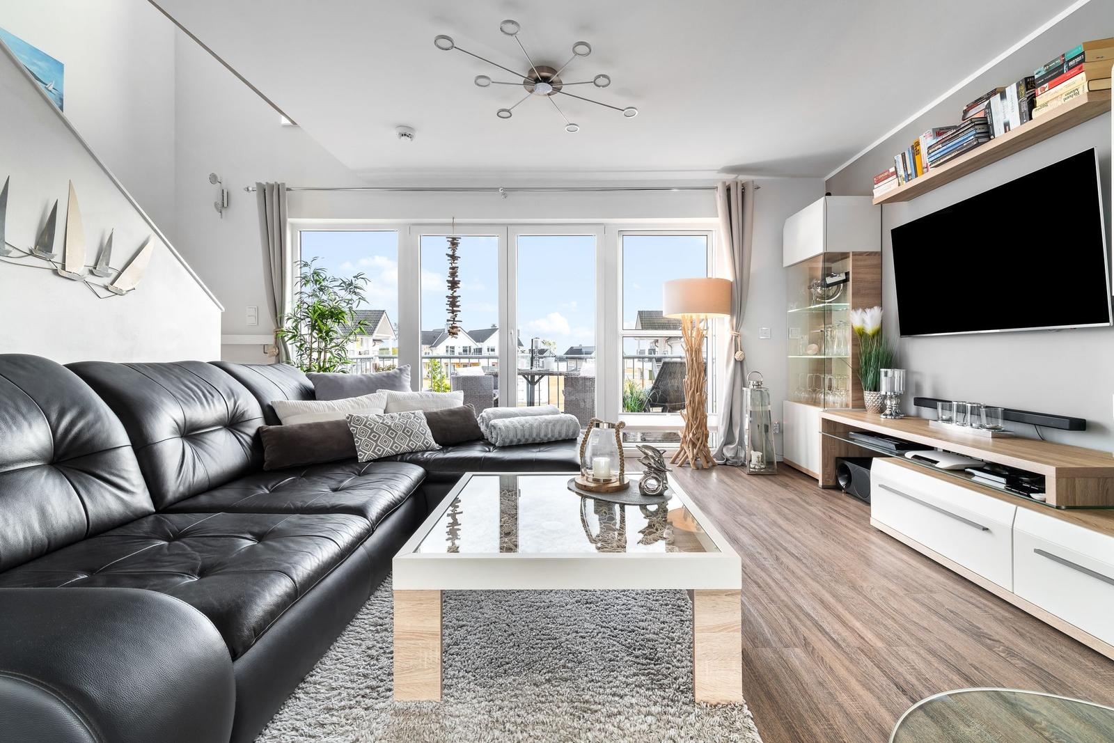 Wohnzimmer mit schwarzer Ledercouch, Glas-Tisch und Fernseher an der Wand.