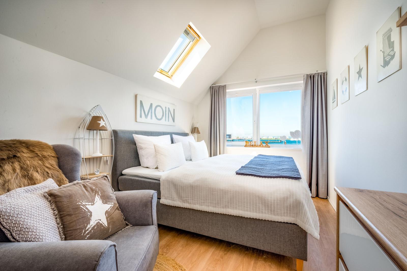 Schlafzimmer mit Dachschräge, Bett, Sessel und Fensterblick auf Wasser.