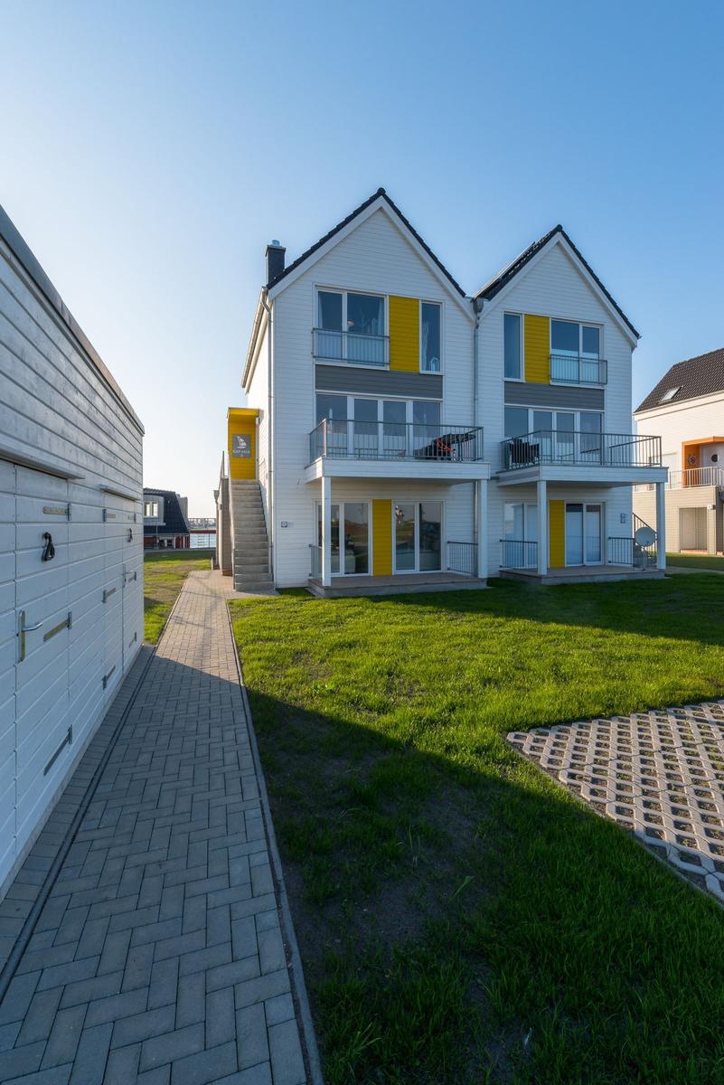 Zwei-Stöckiges Haus mit Balkonen und gelben Akzenten. Grünfläche und gepflasterter Weg.