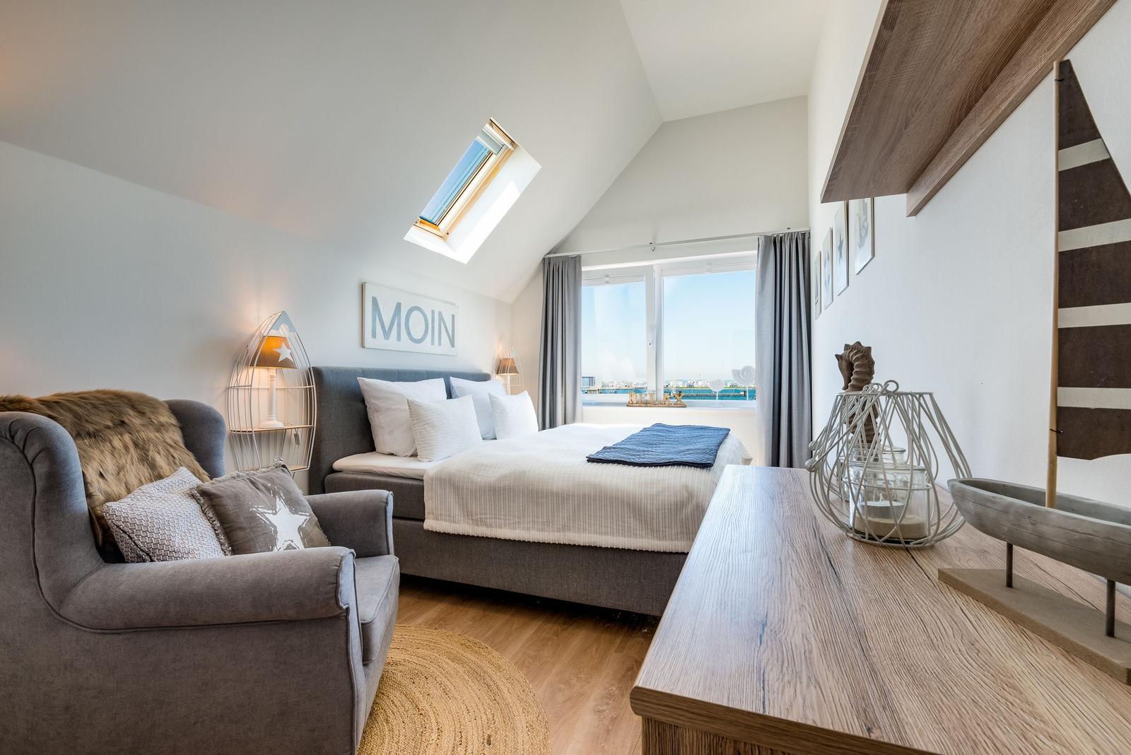 Schlafzimmer mit Dachschrägen, Bett, Sessel und Blick auf Wasser.