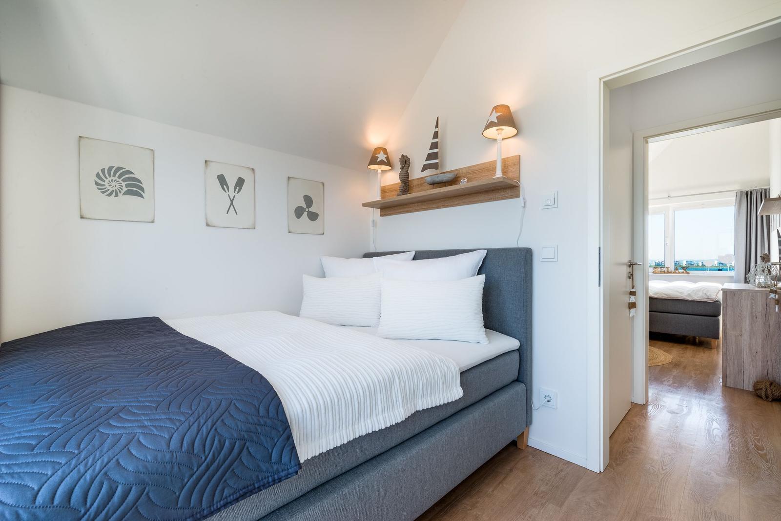 Schlafzimmer mit Bett, Wandbildern und Nachttischen. Durchgang in einen weiteren Raum.