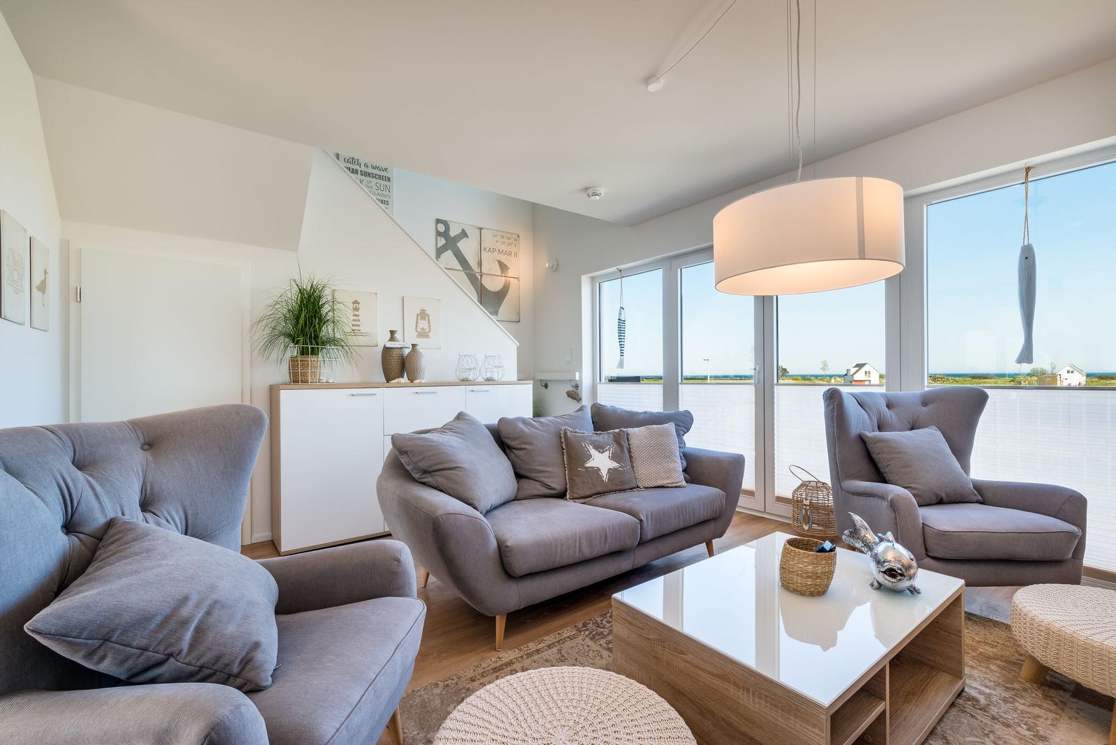 Wohnzimmer mit grauen Sesseln, Couch und Glas-Tisch. Große Fenster mit Meerblick.