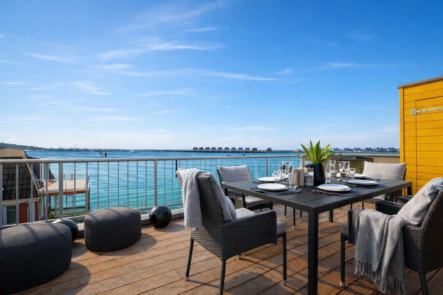 Terrasse mit Esstisch, Stühlen und Meerblick. Holzdeck, Geländer und gelbe Wand sichtbar.