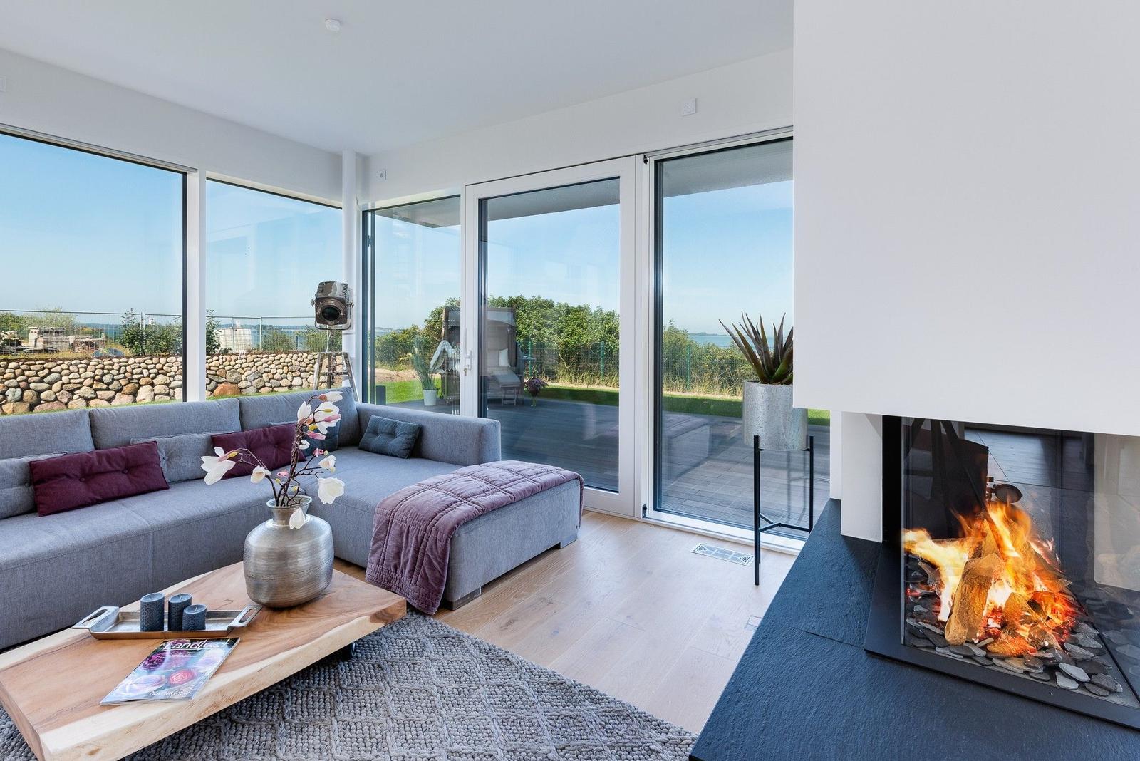 Ein Wohnzimmer mit Sofa, Kamin und großflächigen Fenstern mit Blick auf den Garten.